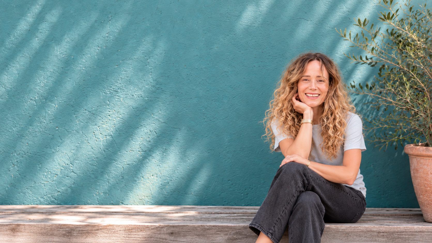 Susannah Saunders smiling, sitting on a wooden step in front of a teal wall
